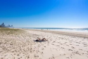 A beach with a person lying on the sand, a city skyline in the background, and a clear blue sky. - 1 & 2 / 39 Albatross Avenue (Gold Coast)