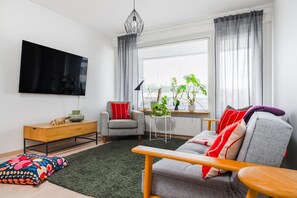 Ett modernt vardagsrum med en platt-TV, en grå soffa, ett träbord och ett litet sidobord med en lampa.