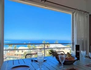 Una mesa dispuesta para comer con vista al océano, un bol de fruta y una botella de vino.