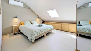 A bedroom with a sloped ceiling, a bed with patterned bedding, a bedside table, a wall-mounted air conditioner, and a ceiling light fixture. - Vrbo Property (Saint-Paul)