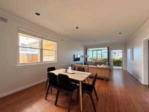 A spacious room with a wooden floor, a dining table with chairs, a sofa, and a television on the wall. - KozyGuru | Manly | Serene Coastal House Retreat (Manly)