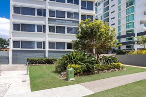 A modern building with a sign reading 'AMBASSADOR' and a well-maintained garden area with various plants and a small tree. - Ambassador Beachfront Burleigh Heads (Gold Coast)
