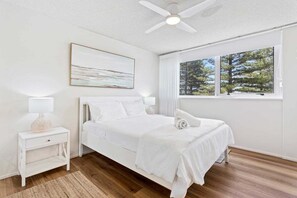 A bedroom with a bed, a nightstand, a ceiling fan, a window with a view of trees, and a painting on the wall. - Ambassador Beachfront Burleigh Heads (Gold Coast)