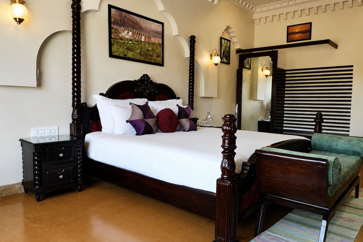 A bedroom with a four-poster bed, a nightstand, a dresser, a mirror, and a view of a mountainous landscape.