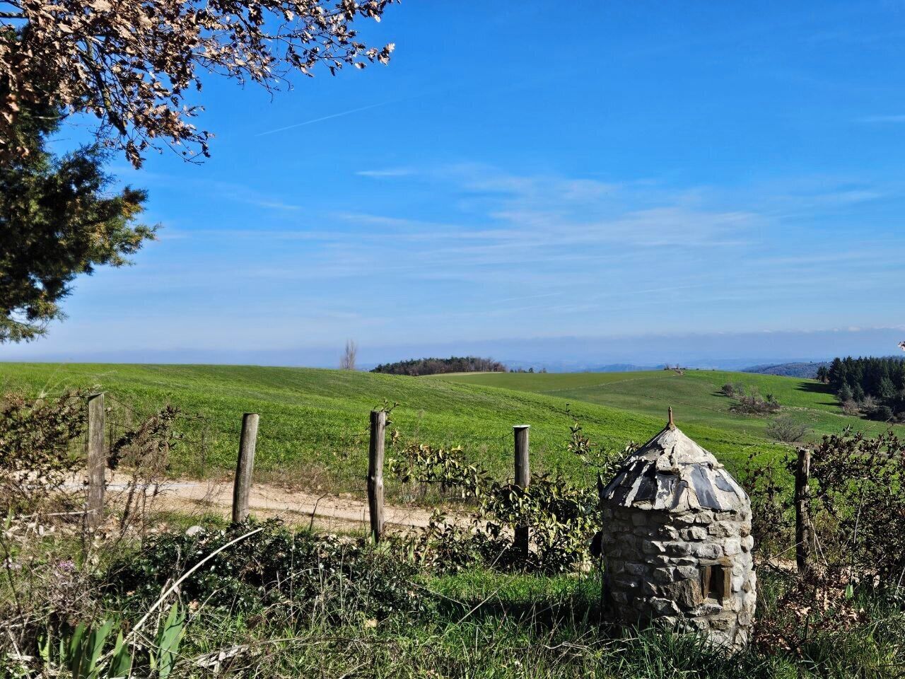 A rural landscape with a stone structure, possibly a well or wellhouse, surrounded by a wooden fence and lush greenery.
