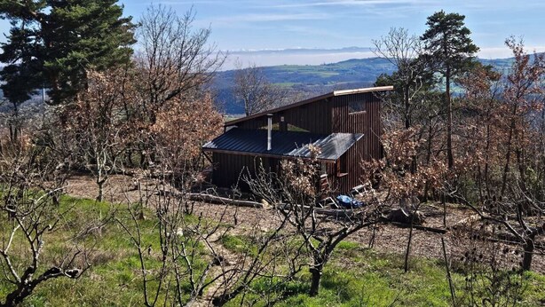Uma cabana rústica com telhado inclinado, rodeada por árvores e uma vista desobstruída de montanhas distantes.