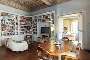 Ein geräumiges Zimmer mit einem Holzespeisetisch, mehreren Bücherregalen, einem Fernseher und einem Kamin.