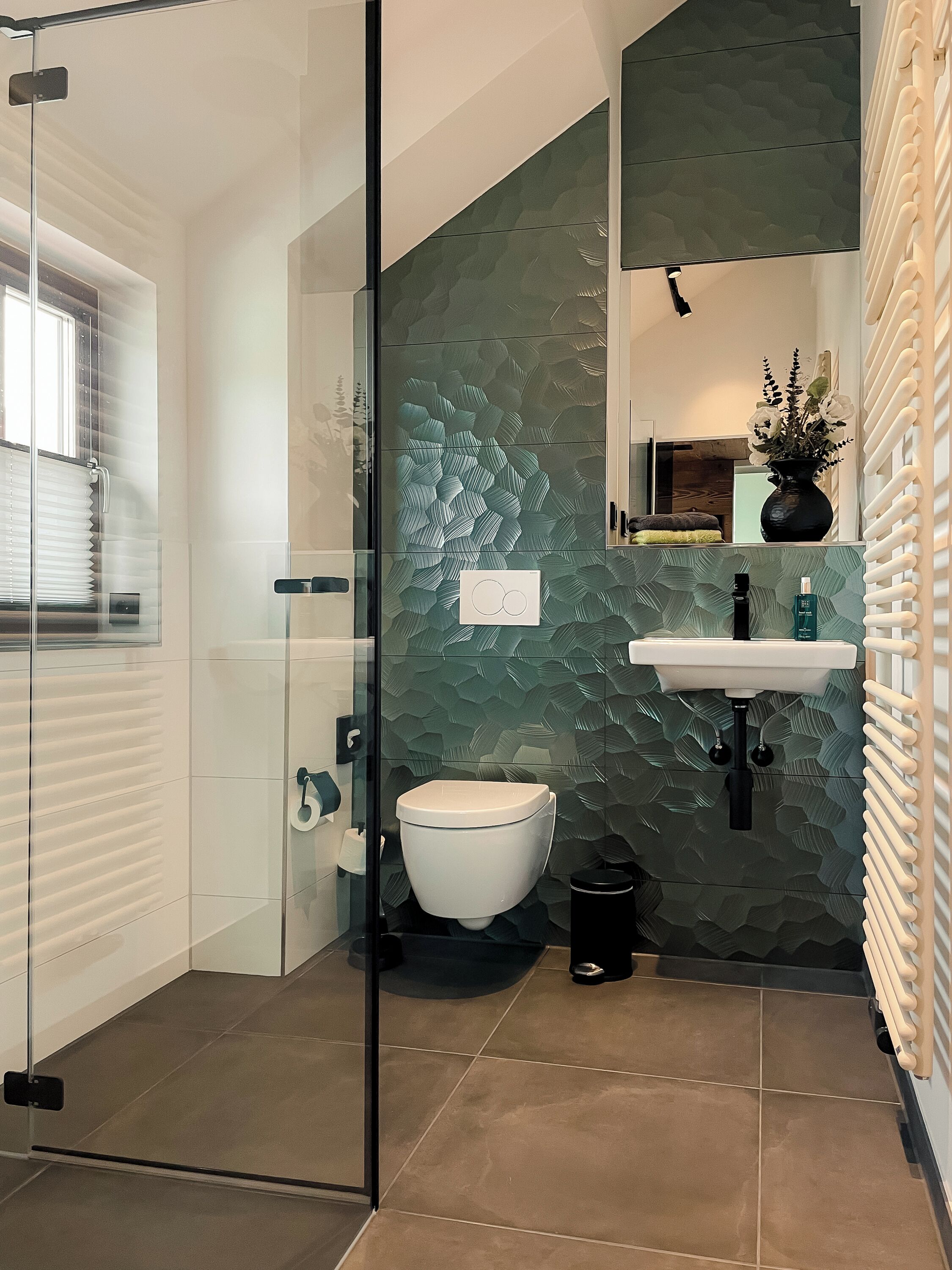 Ein modernes Badezimmer mit Duschkabine aus Glas, einer weißen Toilette und einem Waschbecken, dessen Wand eine grüne Struktur aufweist.
