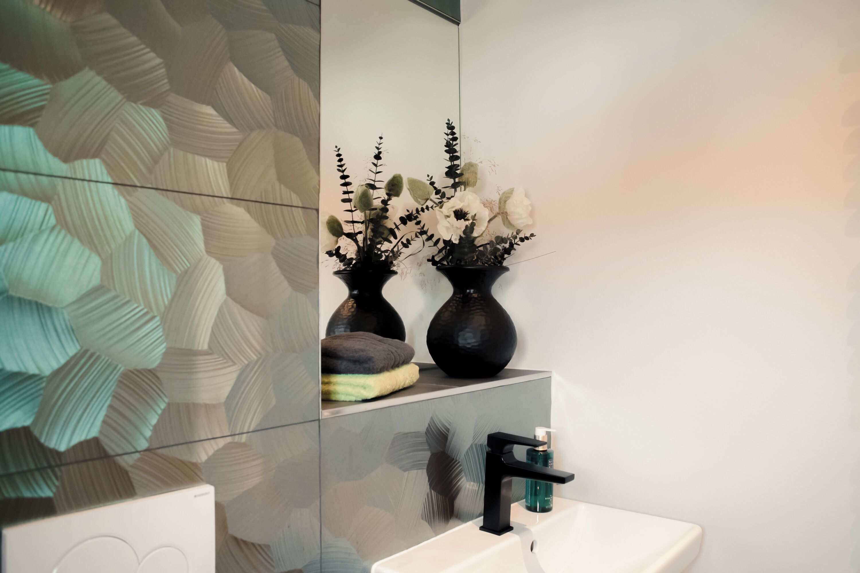 Une salle de bain au design moderne, dotée d’un grand miroir, d’un lavabo blanc et de deux vases noirs ornés de fleurs.