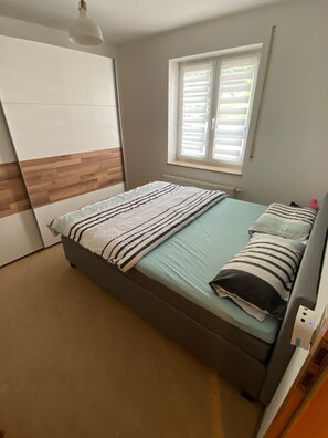 A bedroom with a bed, a window with blinds, a radiator, and a light fixture. - 2-room apartment in green setting near Rheinfelden-Adelhausen, terrace & garden (Rheinfelden)