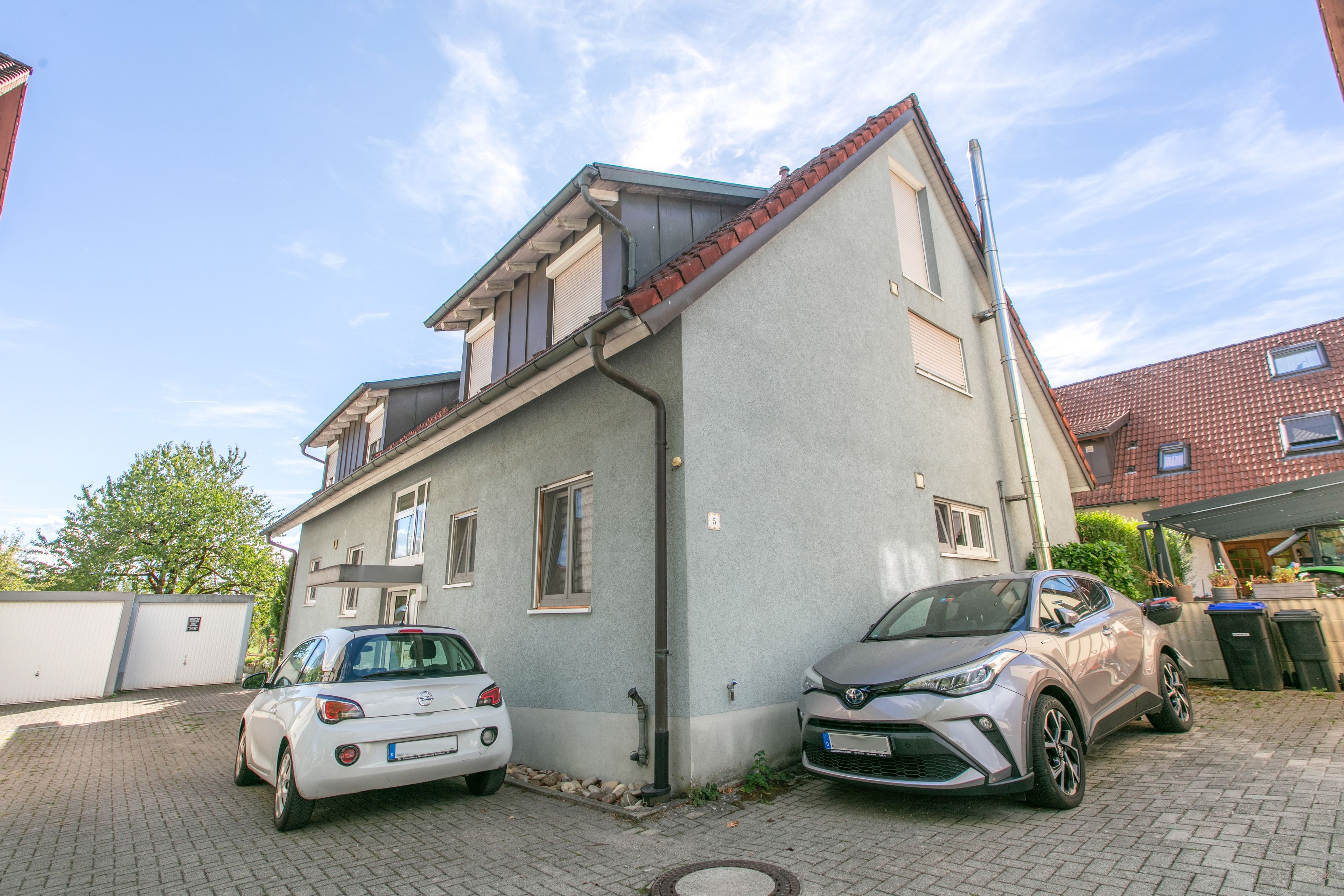 Ein zweistöckiges Gebäude mit einer grauen Fassade, einem roten Ziegeldach und einem gepflasterten Einfahrt.