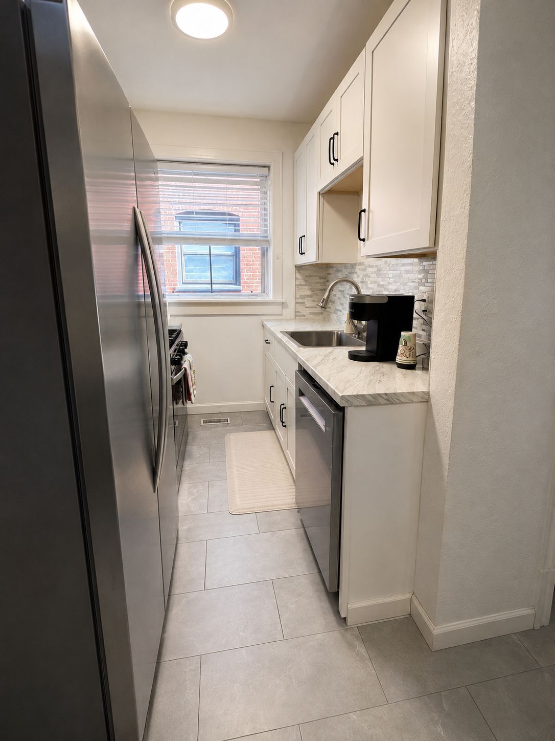 A compact kitchen with a stainless steel refrigerator, a window with blinds, a sink with a faucet, and a countertop with a coffee maker.