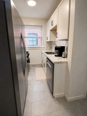 A compact kitchen with a stainless steel refrigerator, a window with blinds, a sink with a faucet, and a countertop with a coffee maker. - Relocate to the Loop • Quiet 2BR near WashU 1N (University City)