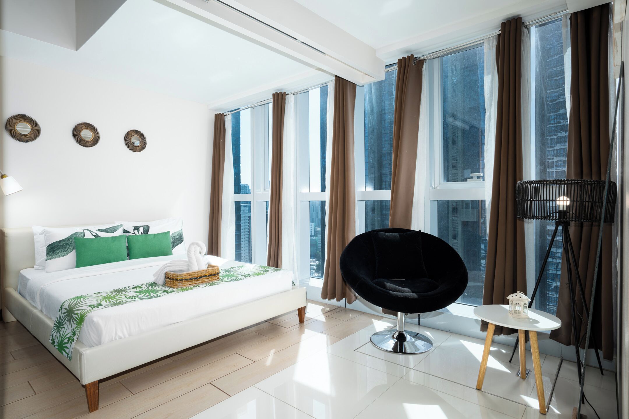 Une chambre d’hôtel moderne avec un grand lit, une chaise noire, une petite table et une vue sur la ville grâce à de grandes fenêtres.