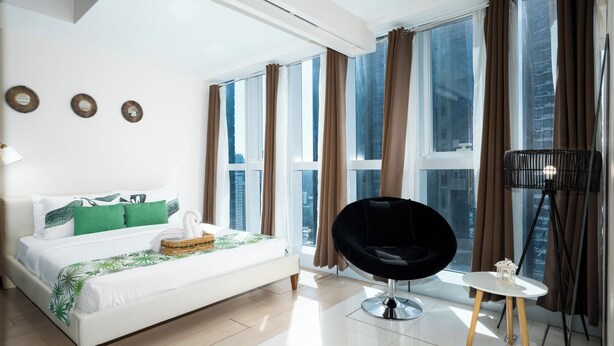 Een moderne hotelkamer met een groot bed, een zwarte stoel, een klein tafeltje en uitzicht op de stad door grote ramen.
