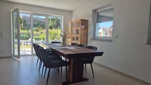 Ein Essbereich mit einem großen Tisch, Stühlen, einer Holzkabine und Blick durch Glastüren auf einen Garten.