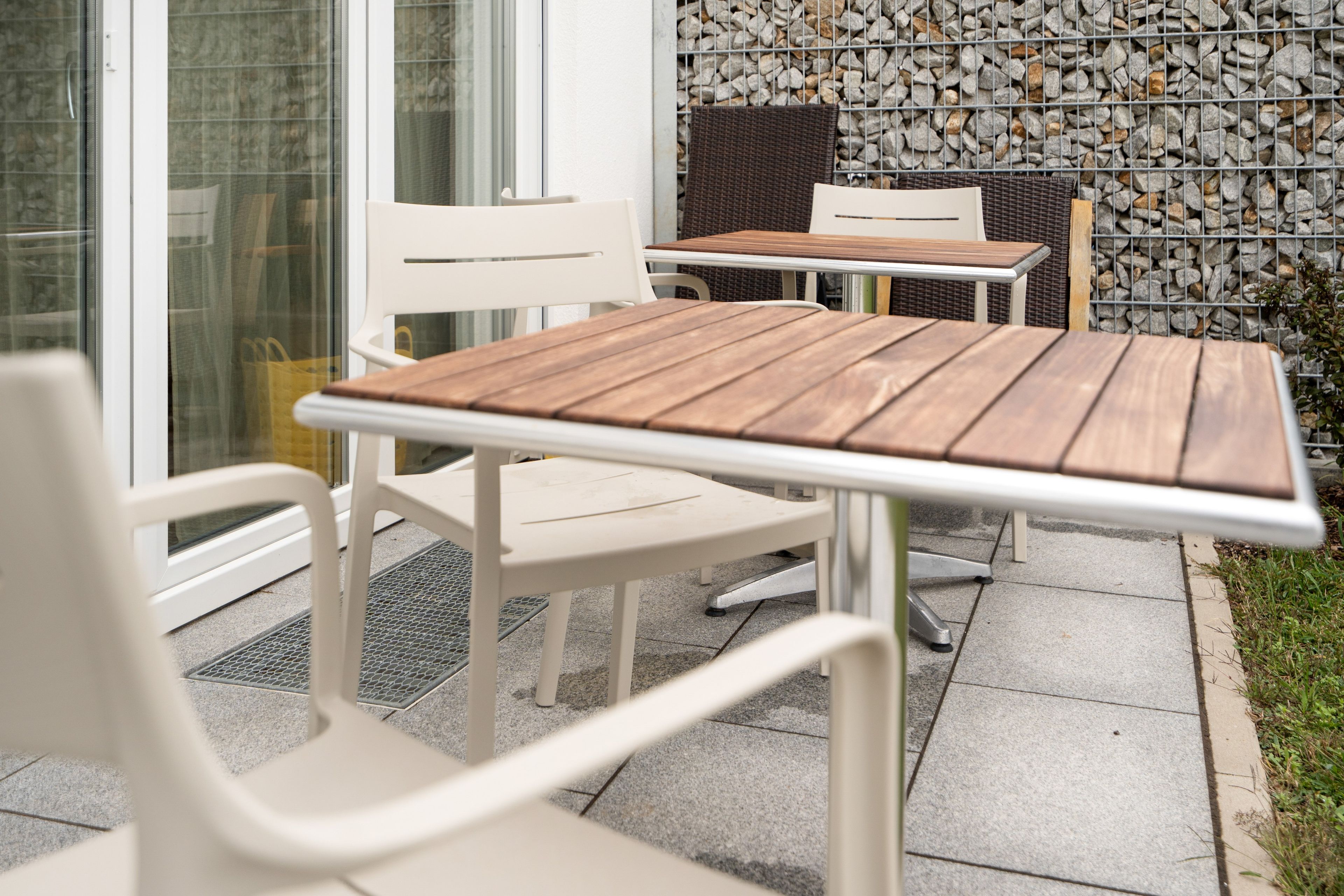An outdoor seating area with white chairs and tables, a stone wall, and a glass door.