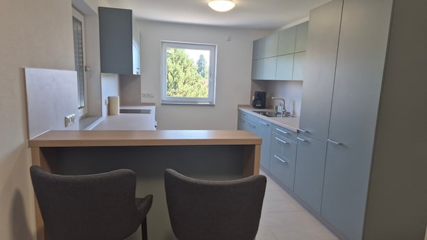 A compact kitchen with a built-in desk and two chairs, a window with a view of trees, and grey cabinets. - Holiday Home 'Gästehaus Klosterblick' with Shared Terrace and Private Garden (Mainburg)