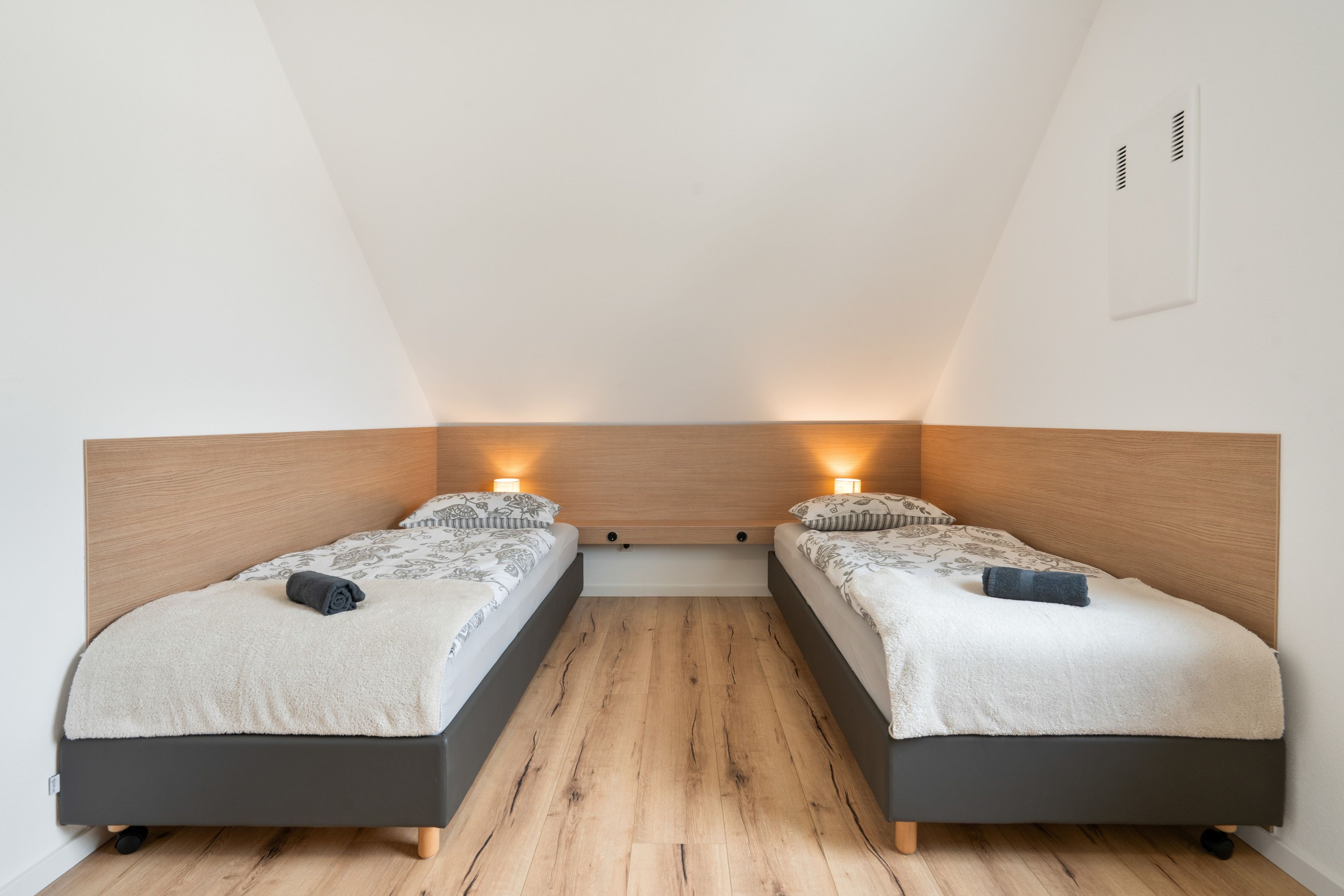 Une chambre avec deux lits, chacun doté d’une tête de lit, d’une table de chevet et d’une lampe. La chambre a un plancher en bois et des murs blancs.