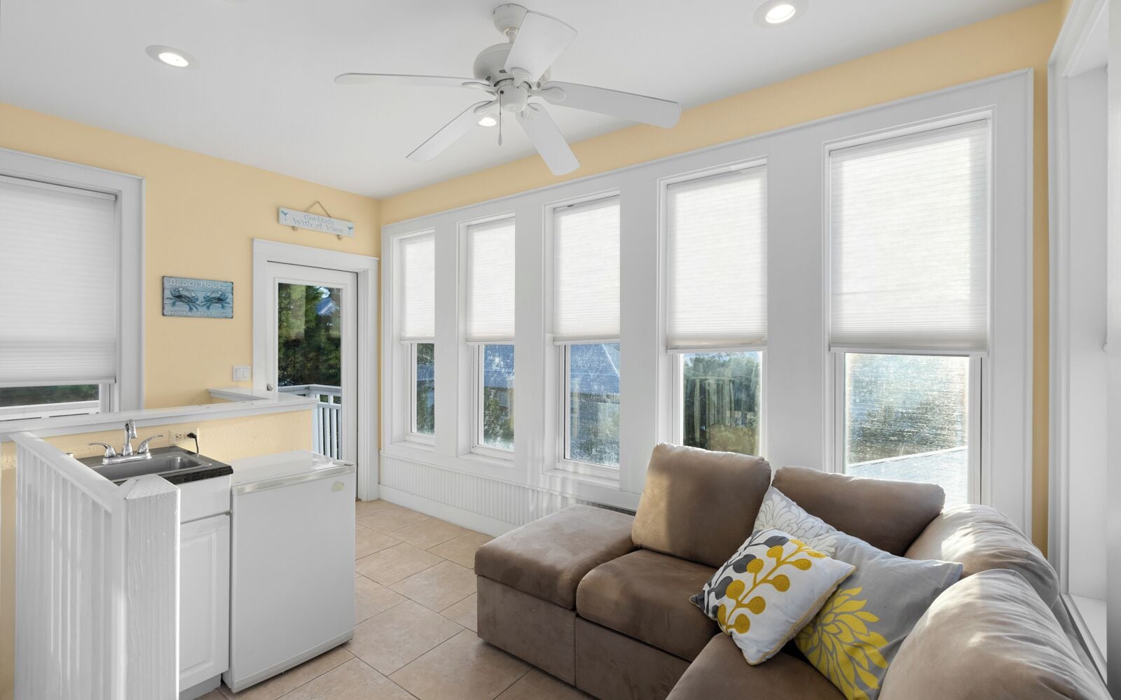 A spacious living area with a kitchenette, large windows, and a ceiling fan.