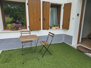 Eine Terrasse mit Holztisch und -stühlen, ein kleiner Gartenbereich mit Kunstrasen und Holzfensterläden.