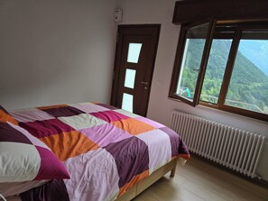 Quarto com cama, janela com vista para paisagem montanhosa, porta e radiador.