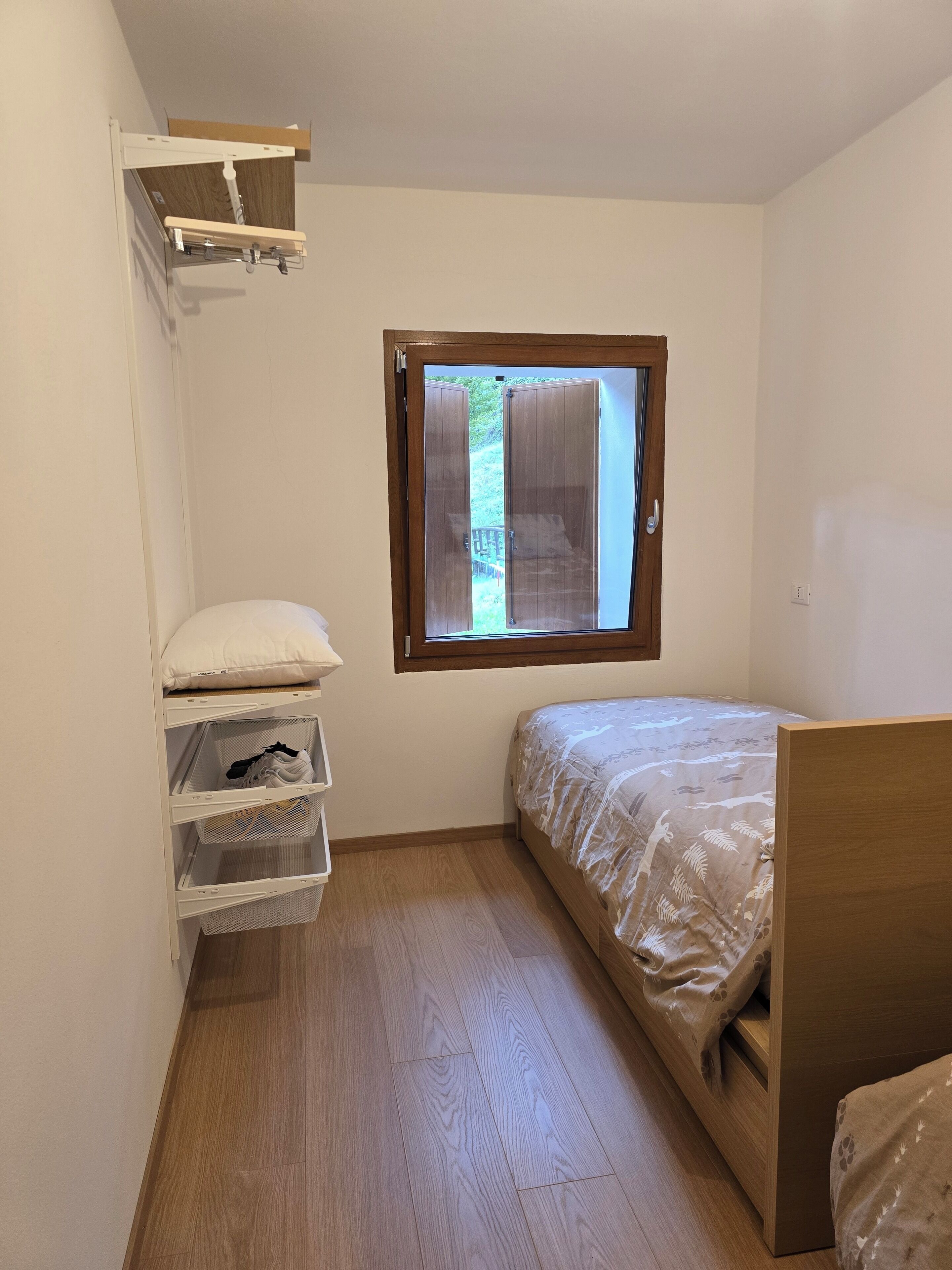 Een compacte, netjes ingerichte slaapkamer met een houten vloer, een bed met een bedovertrek met patroon, een raam met uitzicht op groen en ingebouwde opbergruimte met planken en manden.