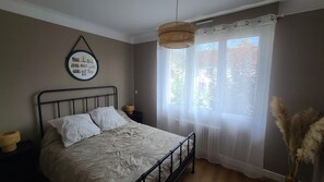 Ett sovrum med en säng, en hängande lampa, ett fönster med tunna gardiner och en tavla som hänger på väggen.