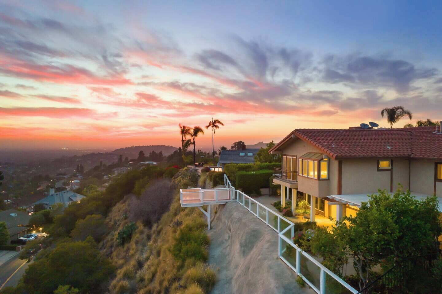 A residential property with a view of the city at sunset, a paved driveway, and a balcony with outdoor lighting.