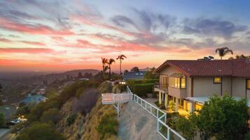 A residential property with a view of the city at sunset, a paved driveway, and a balcony with outdoor lighting.