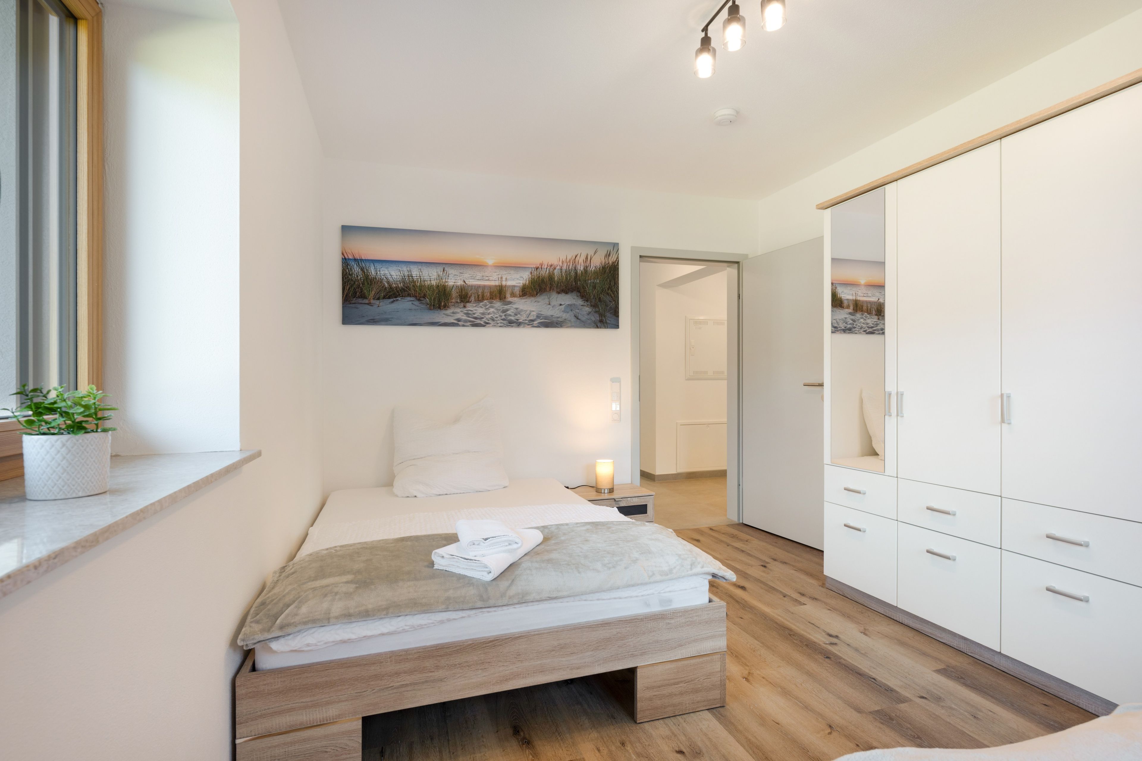 A bedroom with a bed, a nightstand, a bedside lamp, a large wardrobe, and a window with a plant.