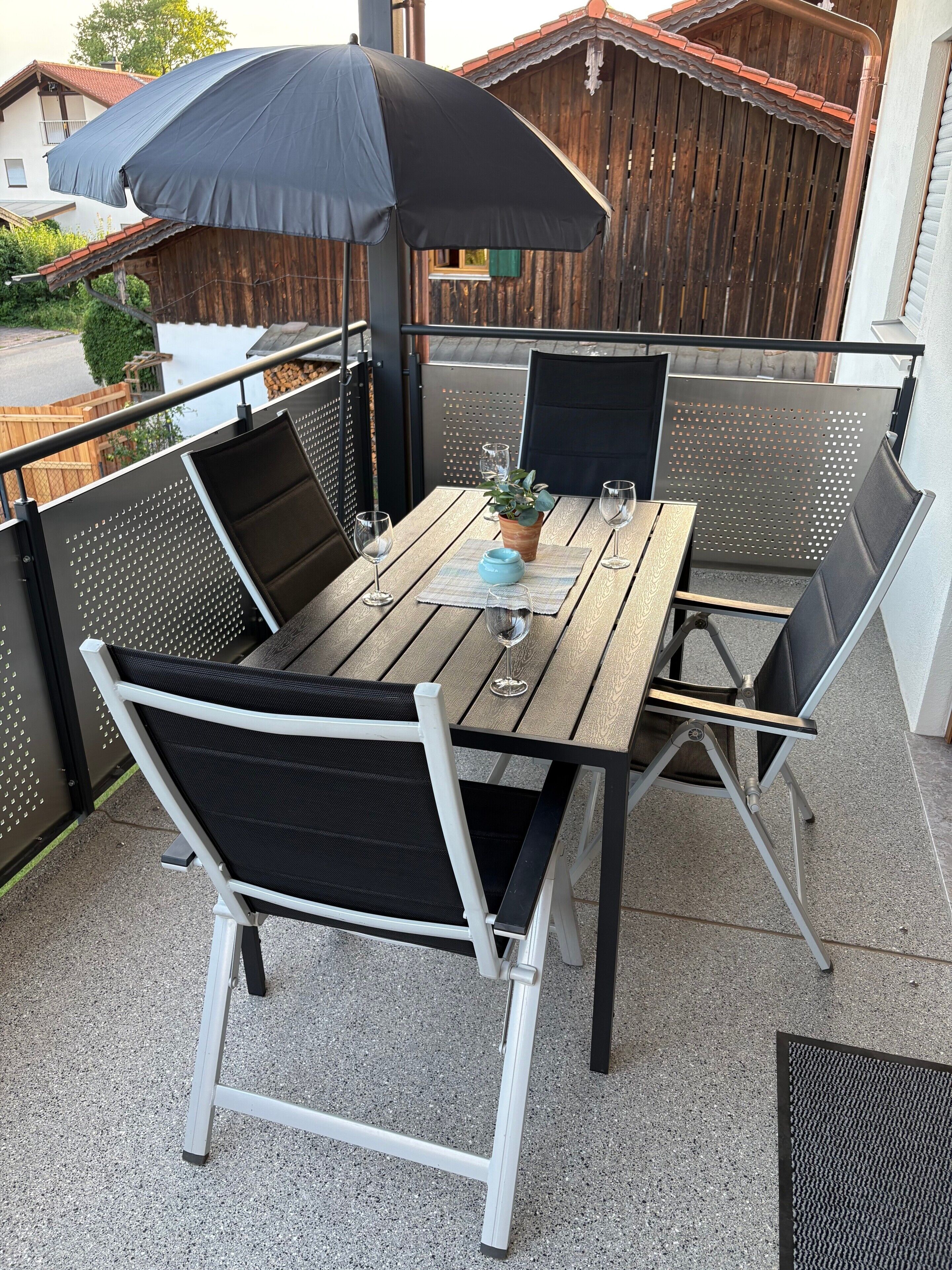An outdoor patio with a wooden table, metal chairs, a black umbrella, and a potted plant.