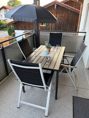 Una terrazza esterna con un tavolo di legno, sedie in metallo, un ombrellone nero e una pianta in vaso.