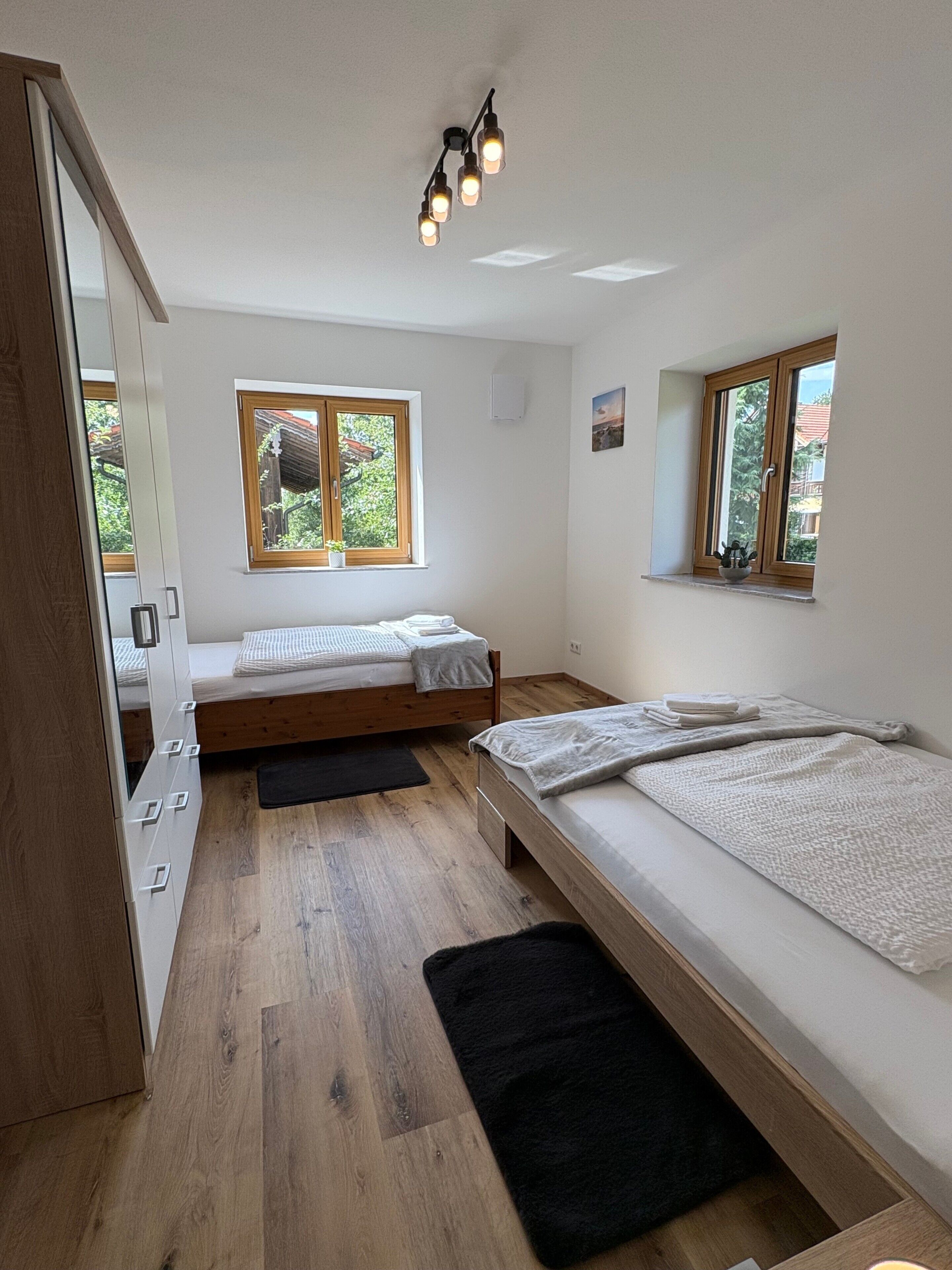 A bedroom with two beds, wooden flooring, a window with a view, and a ceiling light fixture.