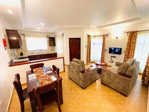 Una sala de estar con un conjunto de sofás, una mesa de comedor con sillas, un televisor y una zona de cocina con armarios y una ventana.