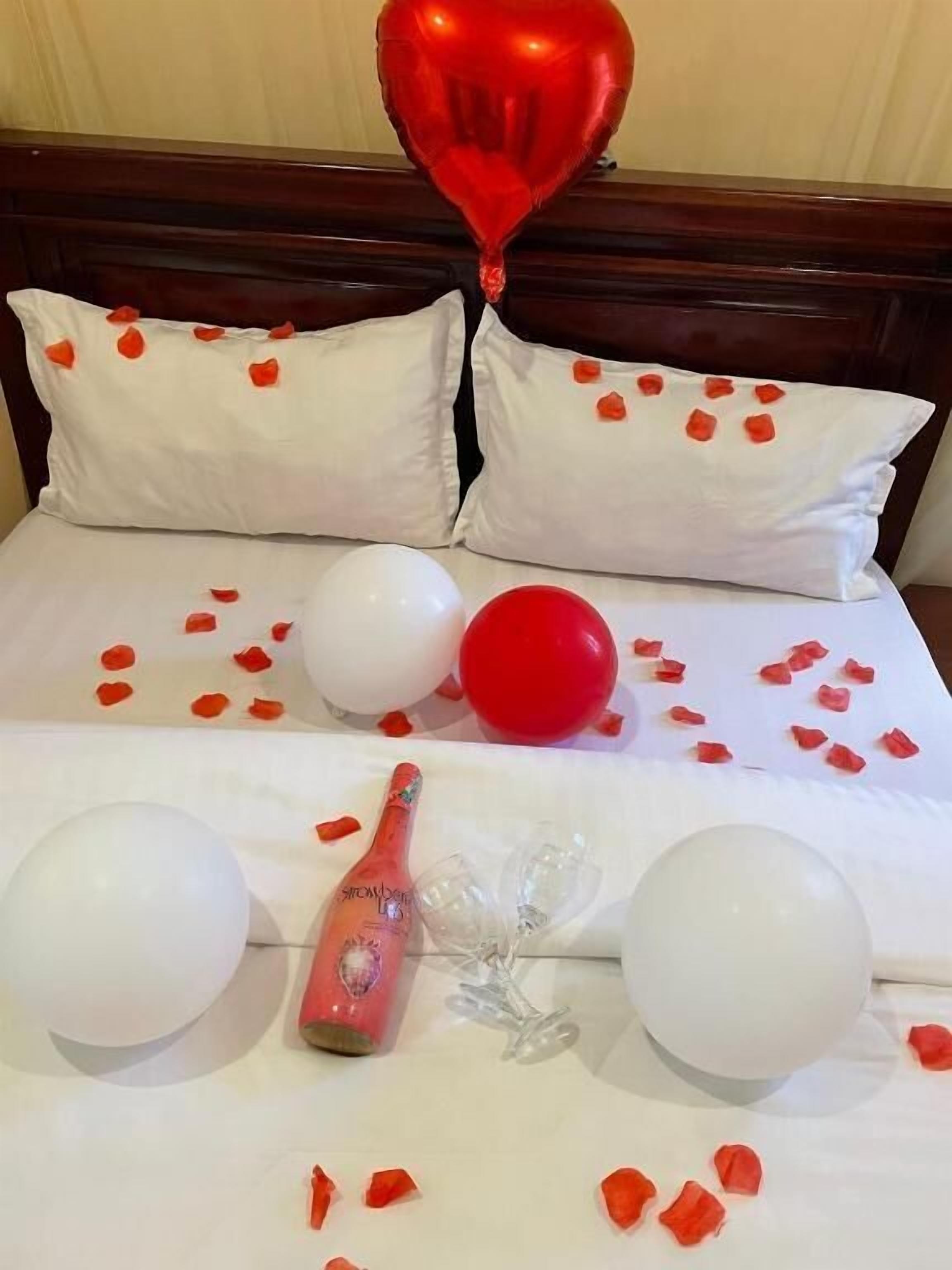 A bed with white pillows, a red heart-shaped balloon, and scattered rose petals.