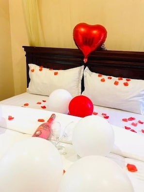 A bed with white bedding, a red heart-shaped balloon, and rose petals scattered on the bed.