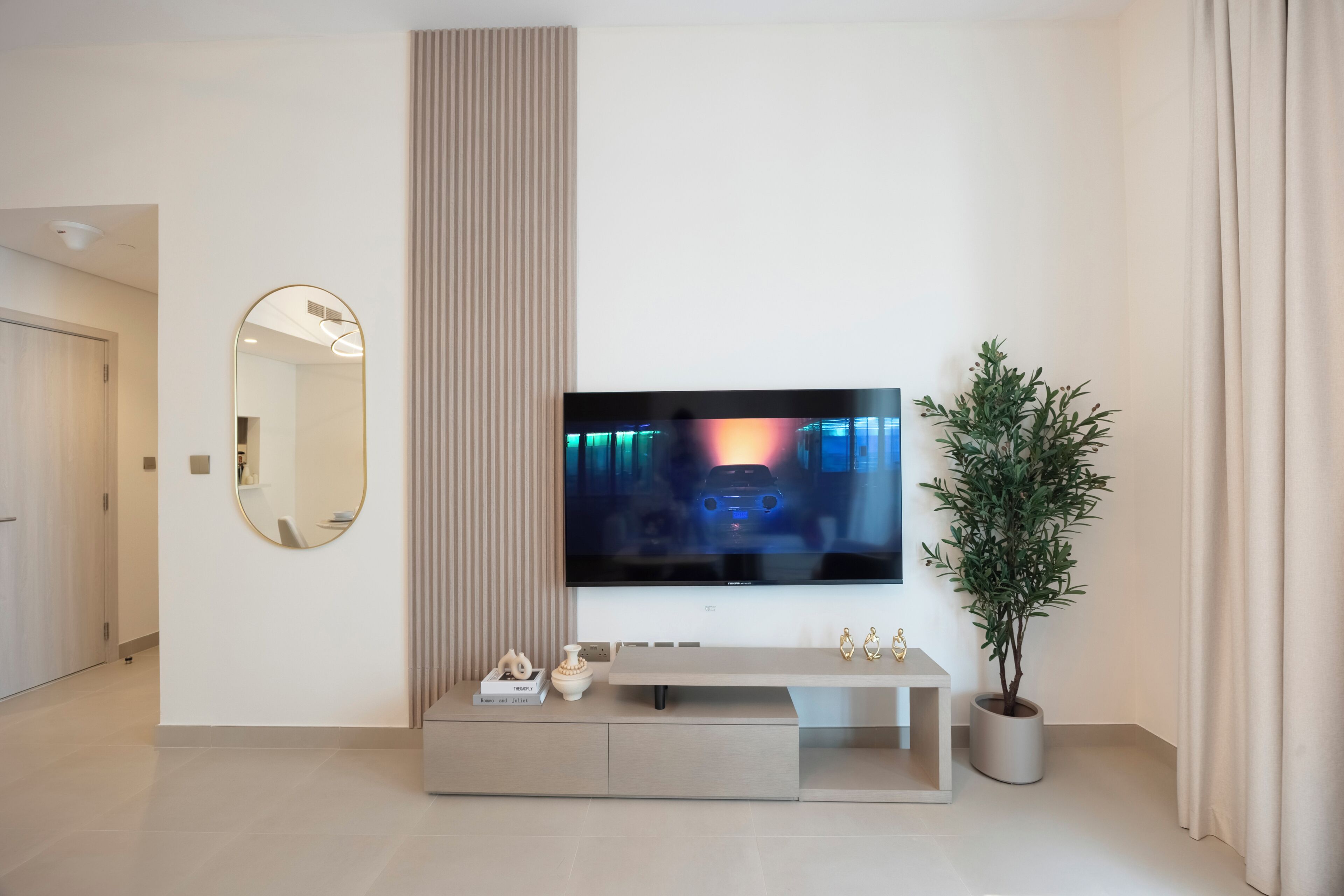 A modern living room with a flat-screen TV mounted on the wall, a round mirror, a potted plant, and a minimalist console table.