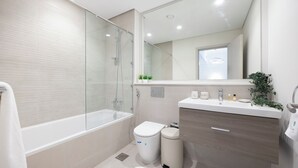 A modern bathroom with a glass-enclosed shower, a white toilet, a trash bin, and a vanity with a mirror. - Bloomfields 1BR in Fairways 1 - Yas (Abu Dhabi)