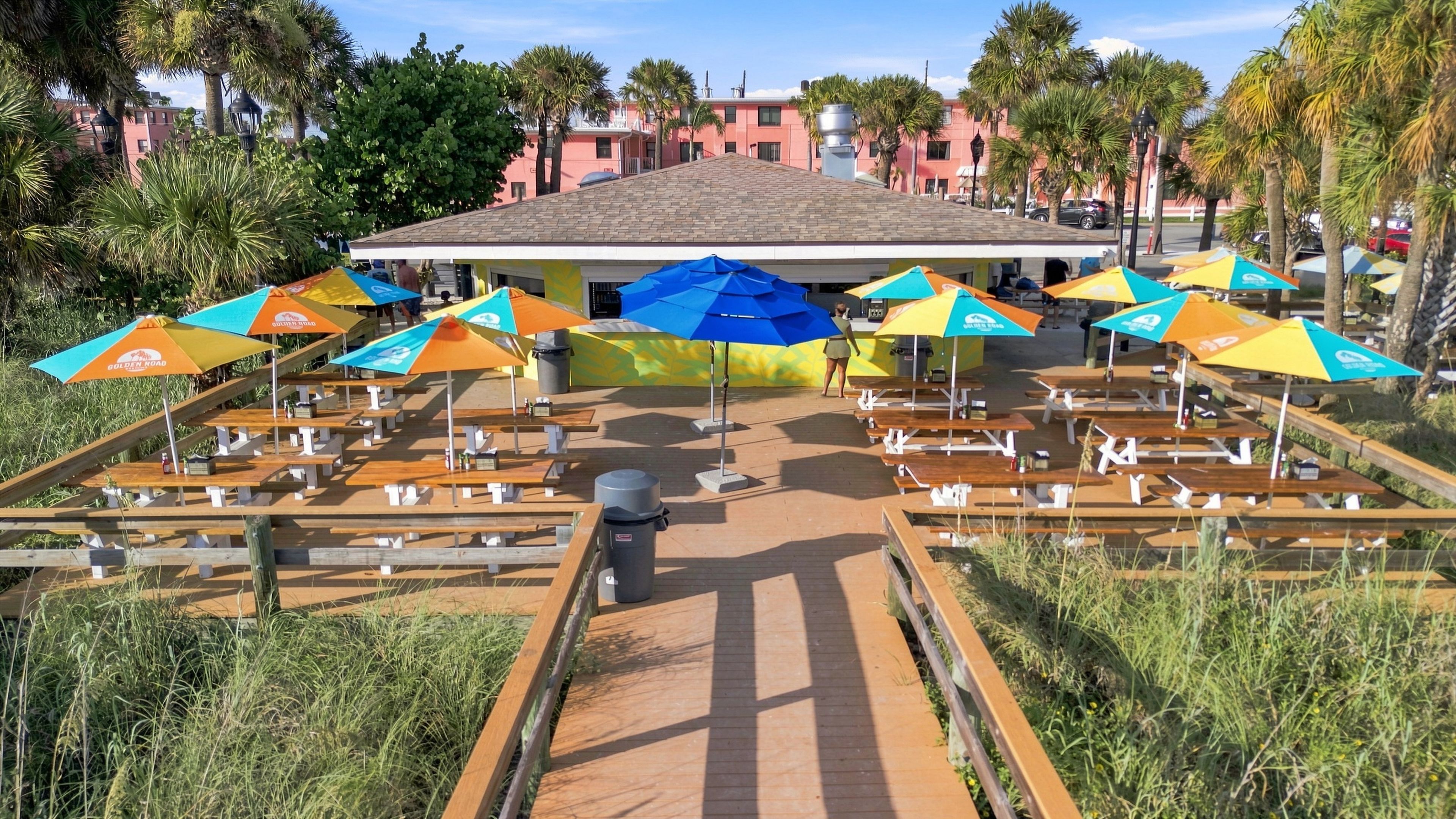 An outdoor seating area with multiple tables and umbrellas, a pathway leading to a building with a tiled roof, and palm trees in the background.