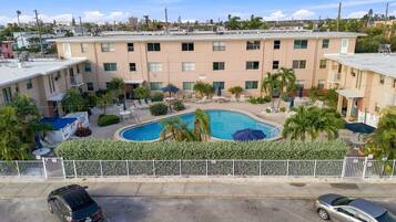 A hotel with a swimming pool surrounded by lounge chairs and umbrellas, a fenced parking area, and a view of the surrounding neighborhood.