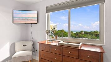 A room with a large window, a television mounted on the wall, a wooden dresser, and a patterned chair.