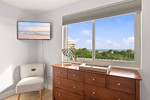 Ein Zimmer mit großem Fenster, einem an der Wand befestigten Fernseher, einem hölzernen Schminktisch und einem gemusterten Sessel.