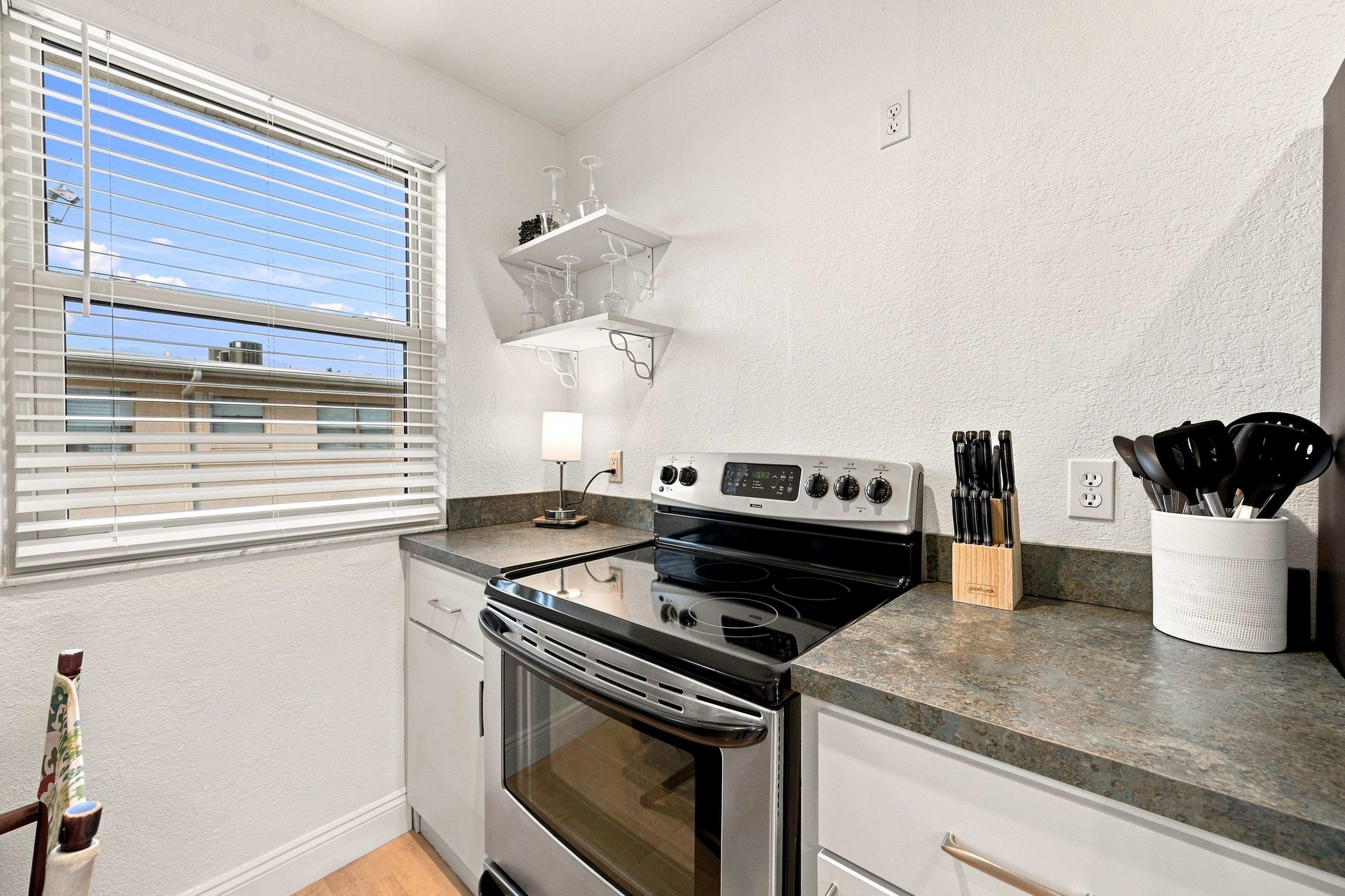 A compact kitchen with a built-in oven, a countertop with a knife holder, and a window with blinds.