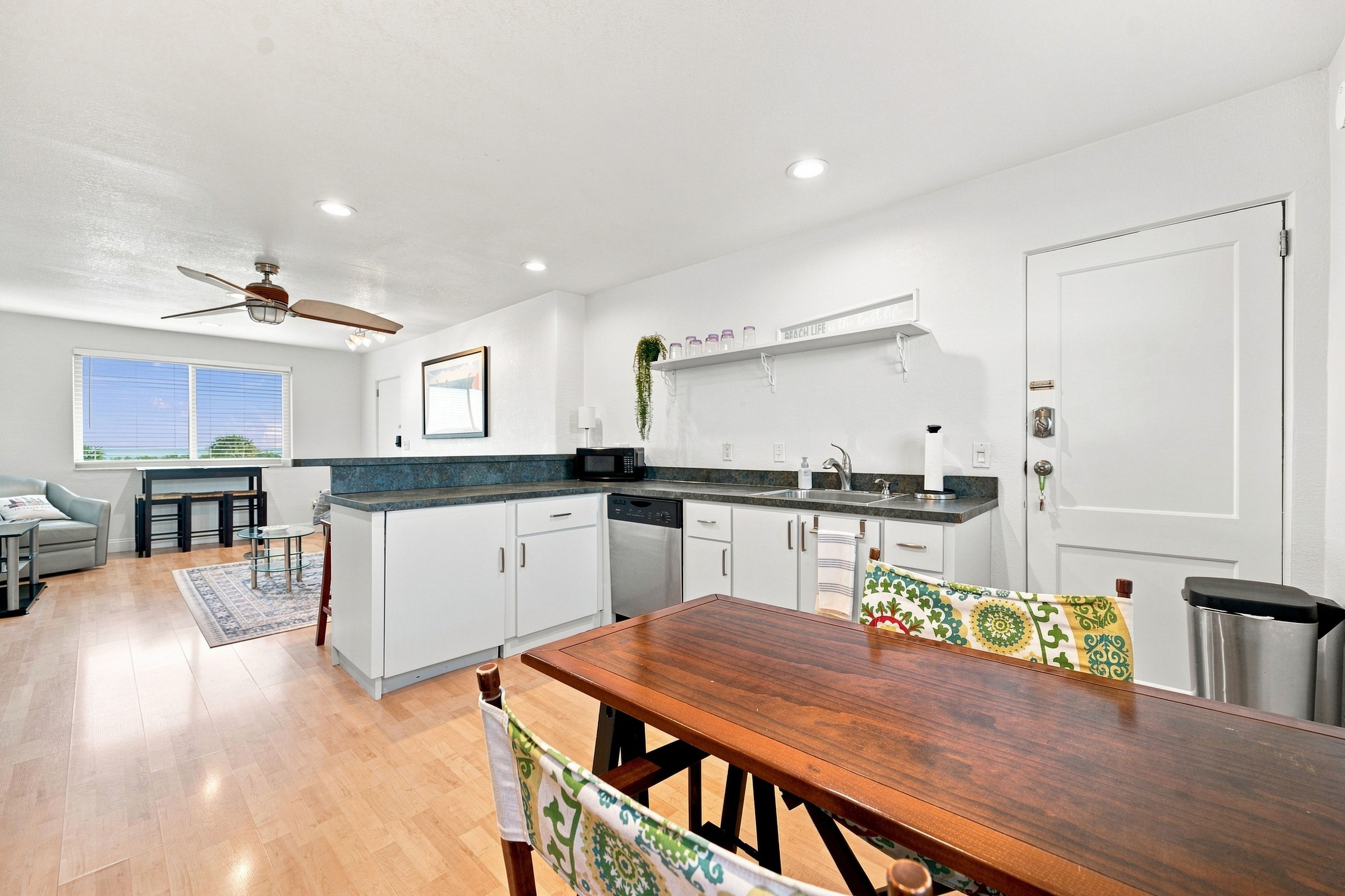 A modern kitchen with white cabinets, a wooden dining table, and a ceiling fan.