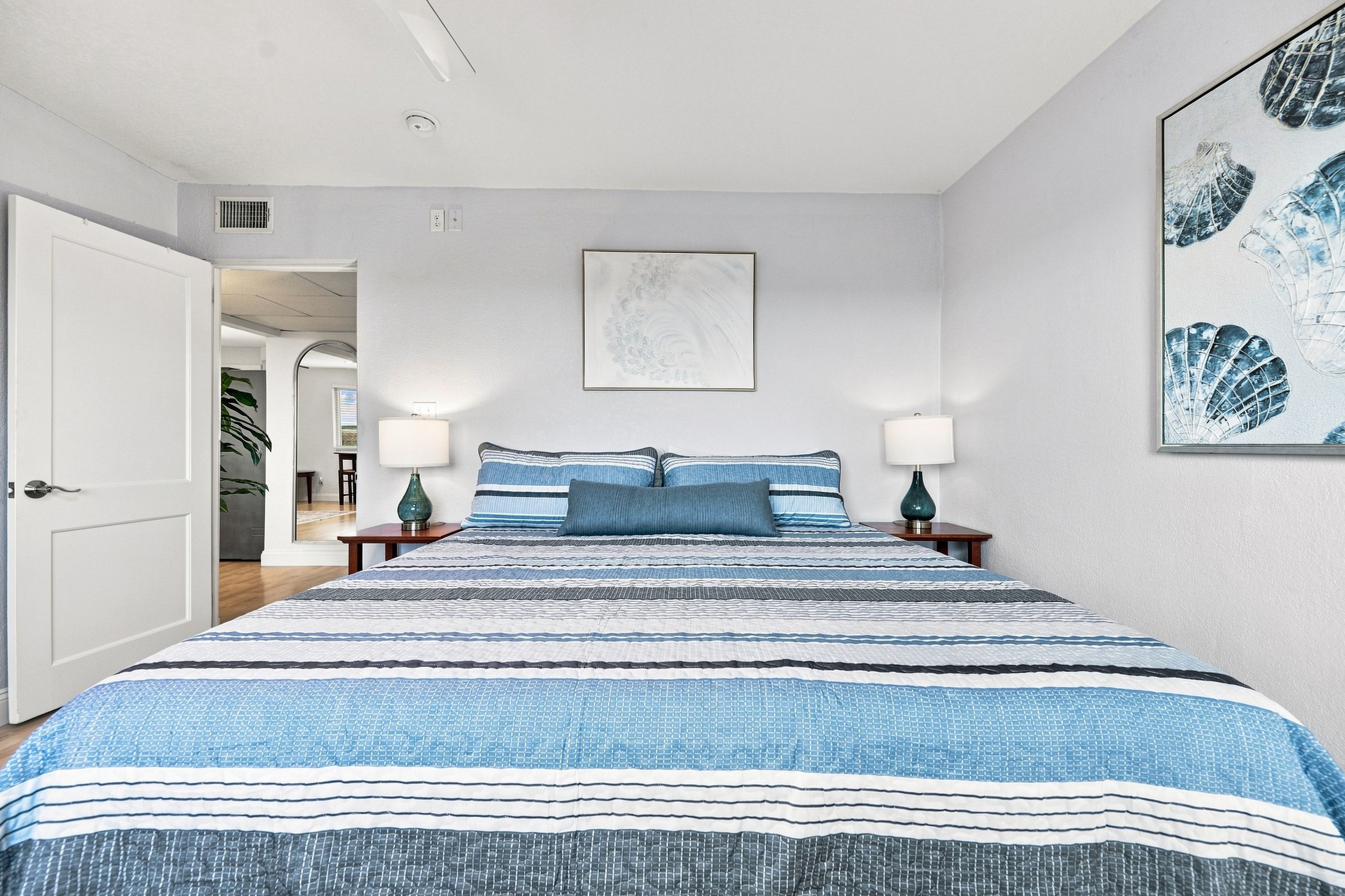 A bedroom with a large bed, two bedside lamps, a framed artwork above the bed, and a door leading to another room.