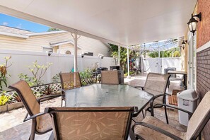 Een terras buiten met een glazen tafel, omringd door rieten stoelen, een wit hek en een bakstenen muur.