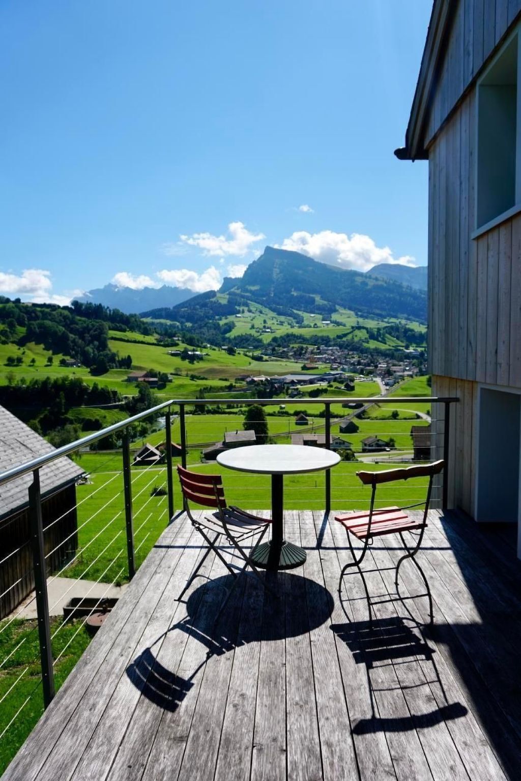 Eine Holzterrasse mit einem runden Tisch und Stühlen, mit Blick auf eine grüne Landschaft und einen Berg in der Ferne.