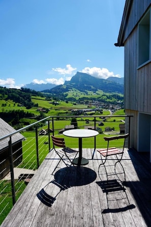 Um deck de madeira com uma mesa redonda e cadeiras, com vista para uma paisagem verde com uma montanha ao longe.