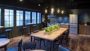 A modern interior with a long wooden table, bar stools, and a kitchen area with a refrigerator. - ALPHABED INN Otaru Ekimae (Otaru)